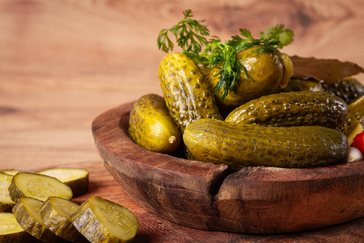 A wooden bowl filled with whole pickles—one of those foods people learn to appreciate later in life—and garnished with dill sits on a table. Sliced pickles are arranged next to the bowl.