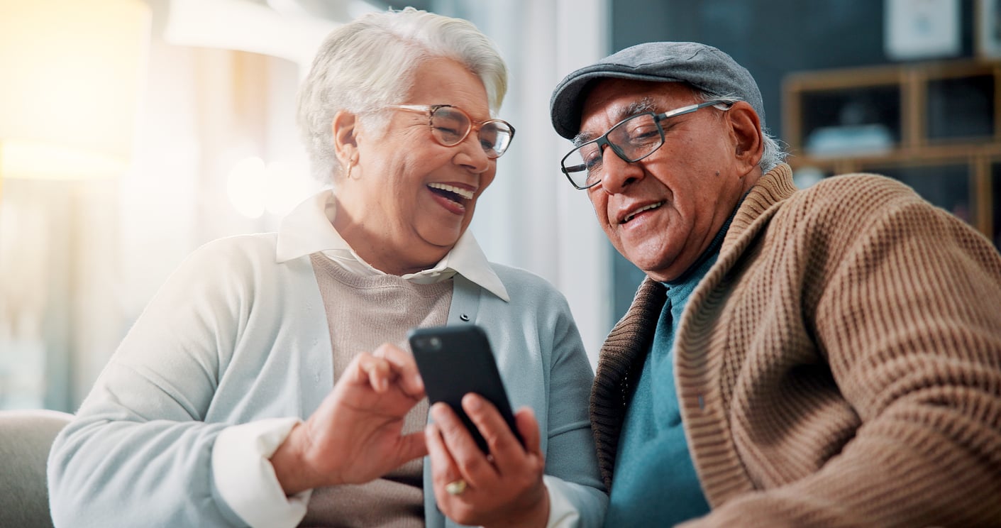 Two older adults sit together on a couch, smiling and laughing while looking at a smartphone. One person is holding the phone and pointing at the screen, while the other wears glasses and a cap. As they enjoy this warm and joyful moment, they’re reminded of how technology has become one of those things families spend less on—bringing loved ones together without breaking the bank.