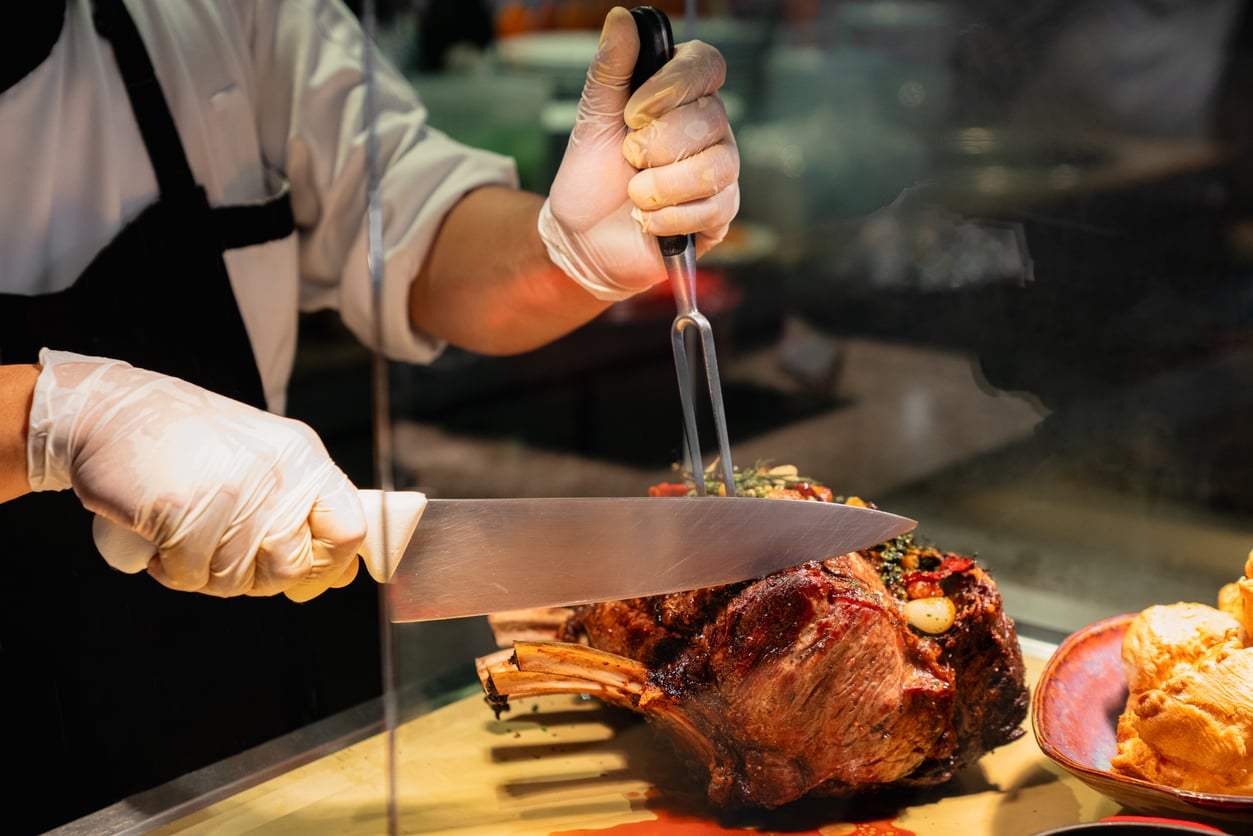 A chef wearing gloves slices a large, roasted rib of meat on a cutting board using a carving fork and knife.