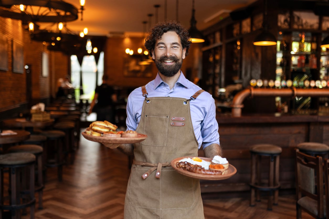 A smiling man with a beard, wearing an apron, holds plates of food in a cozy, warmly lit restaurant with wooden floors and a bar in the background.