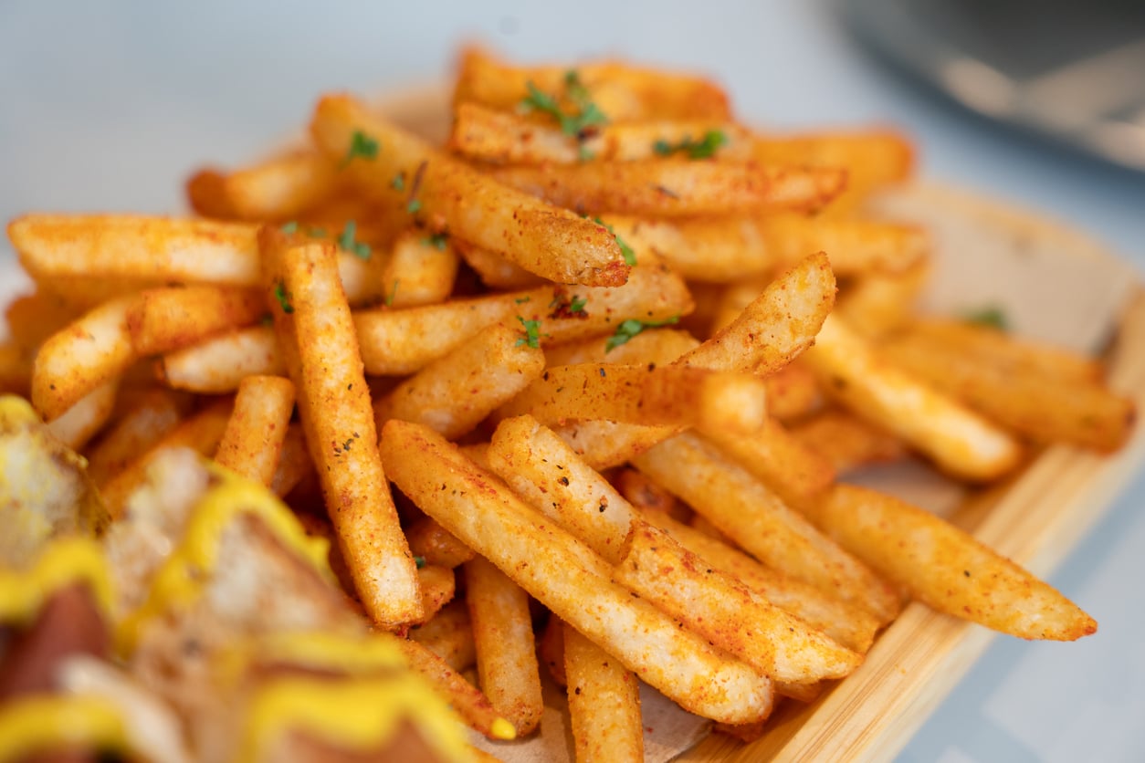 A close-up of seasoned French fries garnished with chopped herbs, served on a wooden tray with part of a sandwich visible in the corner.