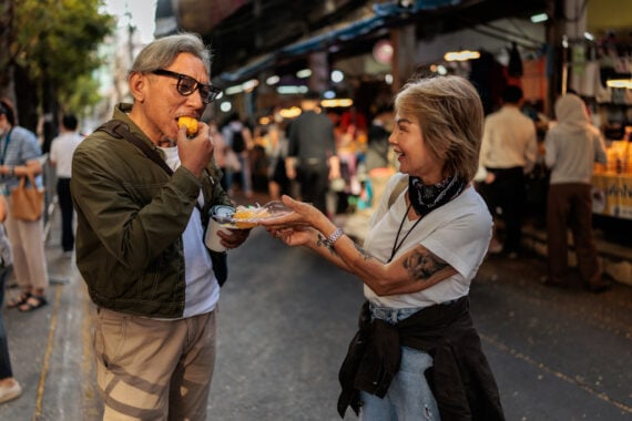 Two older adults smile as one feeds the other a bite of street food at an outdoor market, surrounded by people and vendors&mdash;a heartwarming scene that captures the joy of shared vacation experiences.