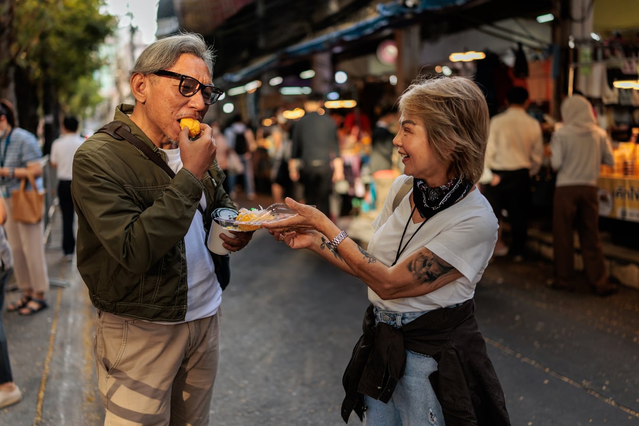 Two older adults smile as one feeds the other a bite of street food at an outdoor market, surrounded by people and vendors—a heartwarming scene that captures the joy of shared vacation experiences.