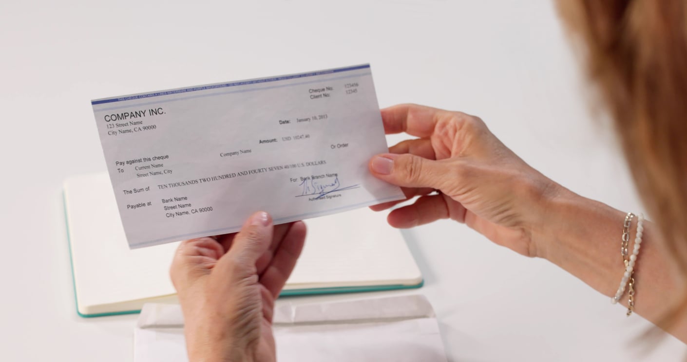 A person holding a check in both hands, preparing to deposit or review it. The check is issued by a company and the background shows a notebook on a white desk.