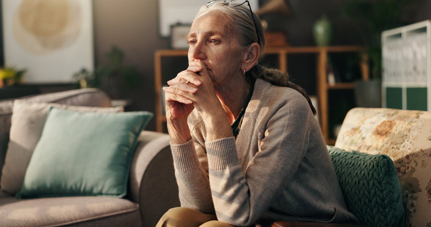 An older woman with glasses on her head sits on a couch with her hands clasped, looking thoughtful—perhaps reflecting on her retirement money habits. The living room has soft lighting, cushions, and decor in the background.