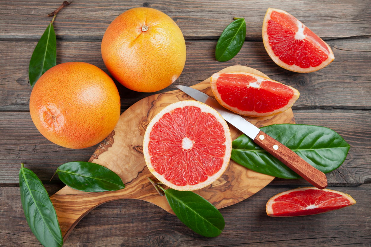 Whole and sliced grapefruits on a wooden board, with a knife and green leaves, placed on a rustic wooden table—a perfect example of the Foods People Learn to Appreciate Later in Life.