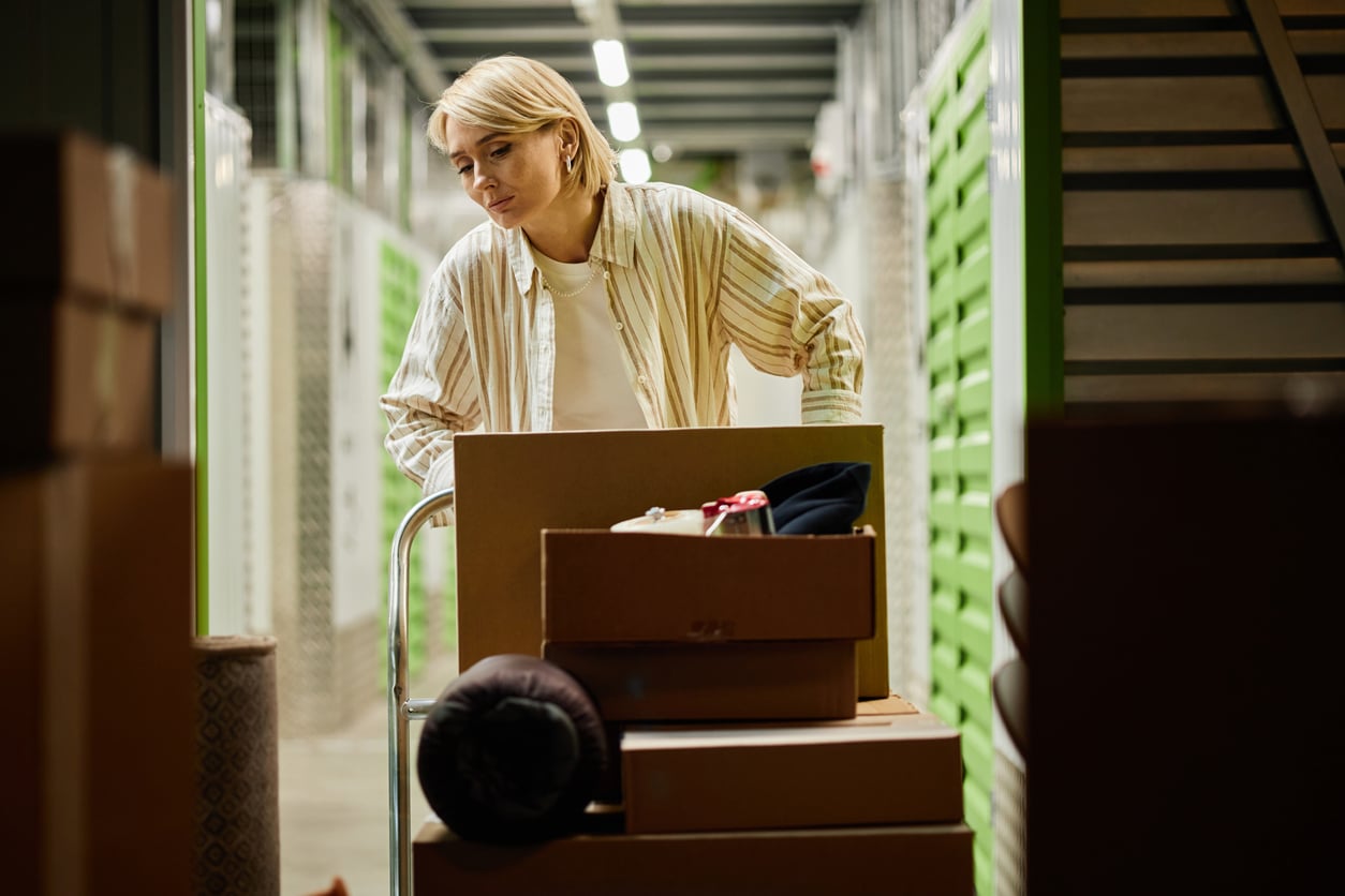 A person with blonde hair pushes a cart loaded with cardboard boxes in a storage facility with green doors visible along the hallway.