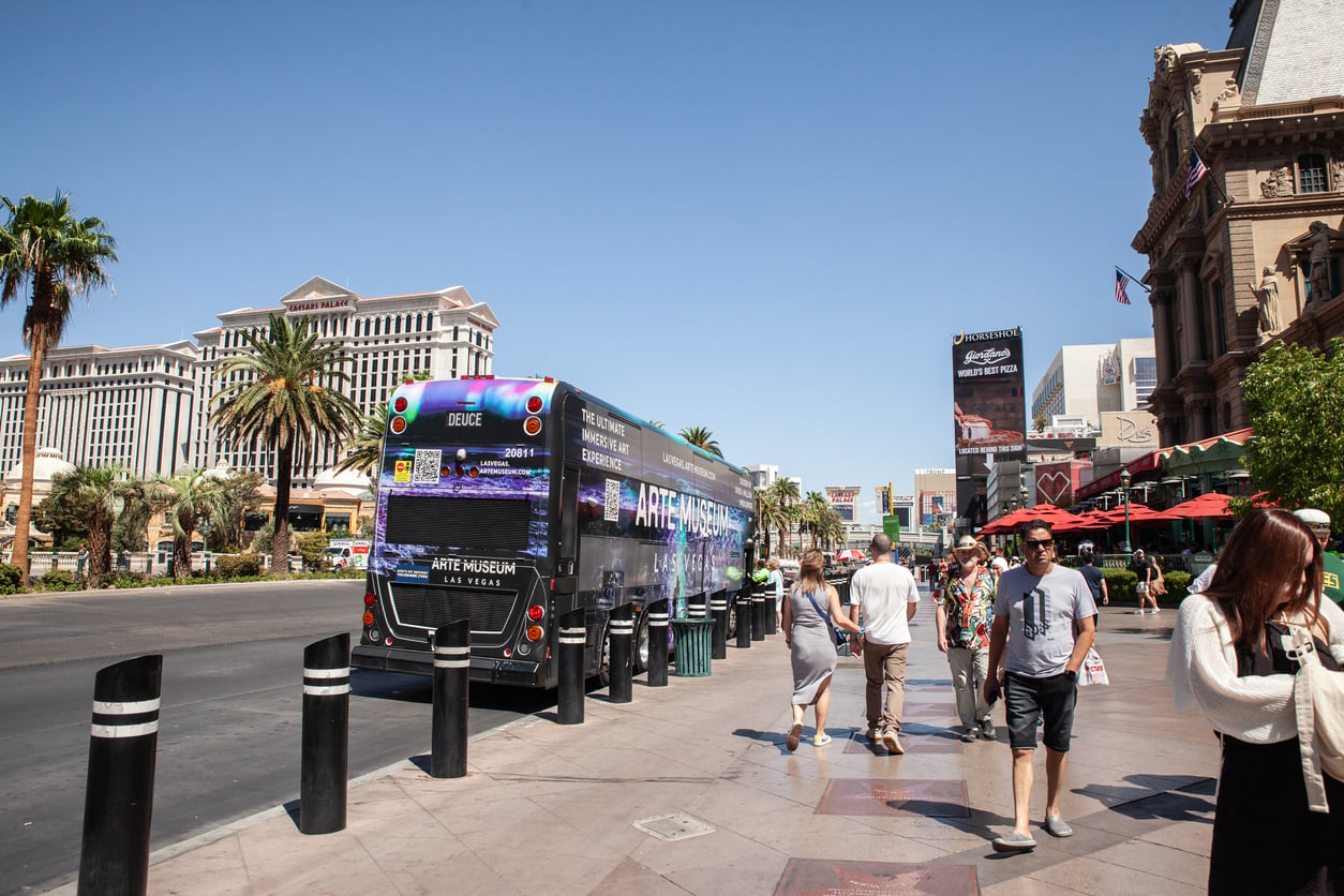 A city street in Las Vegas with a colorful bus parked by the sidewalk, people walking, palm trees, and iconic hotels and signs in the background on a sunny day.