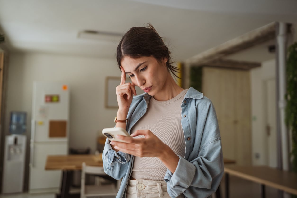 A woman stands indoors, looking at her phone with a concerned expression and touching her forehead, appearing worried or deep in thought. The background shows a kitchen and dining area.