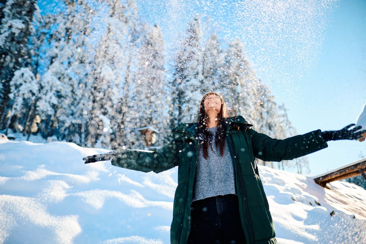 A person in a green coat stands in a snowy landscape, arms outstretched and smiling as snow falls. Pine trees and a clear blue sky are in the background.