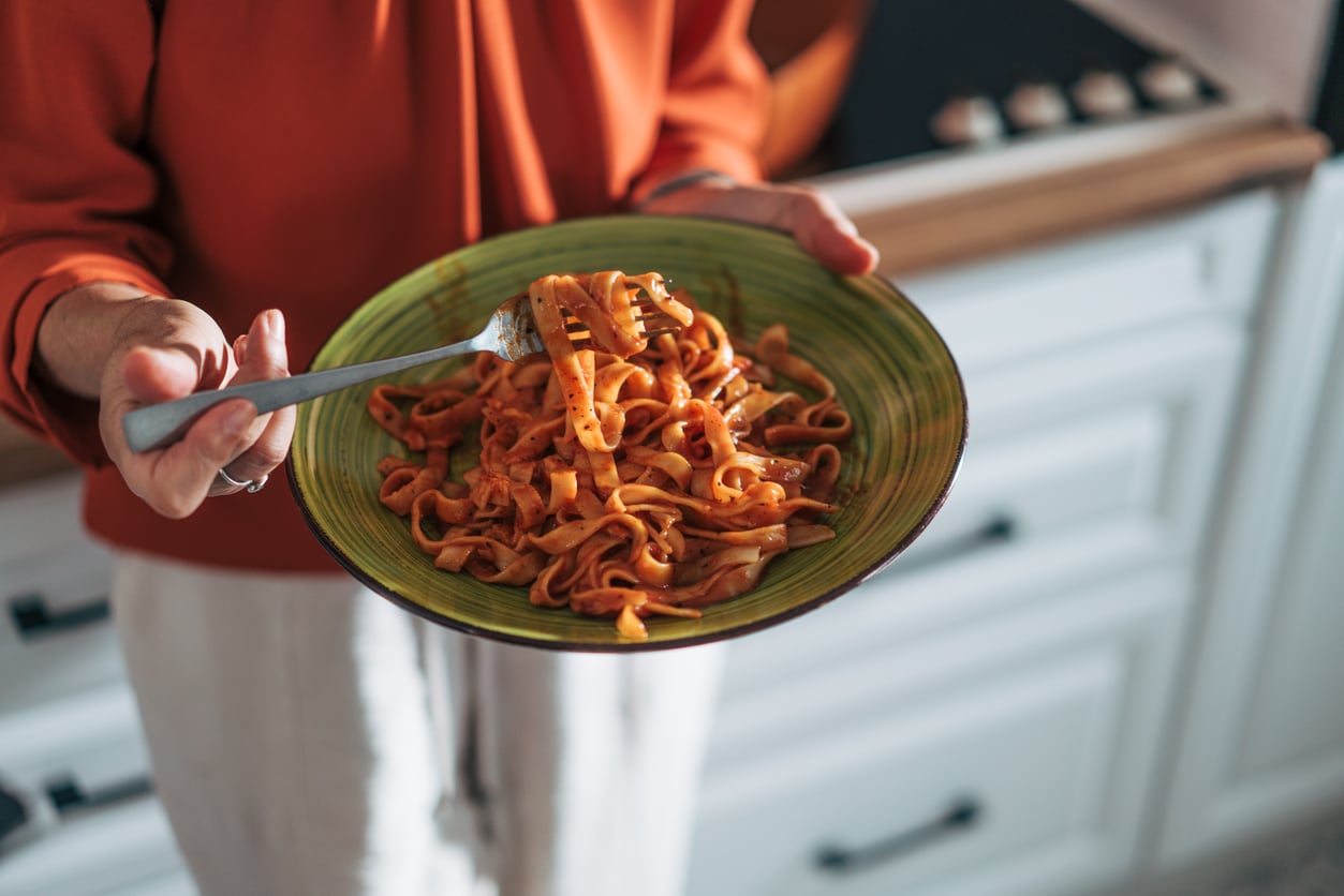 A person in an orange top holds a green plate of pasta with red sauce in one hand and a fork twirling pasta in the other. White cabinets are visible in the background.