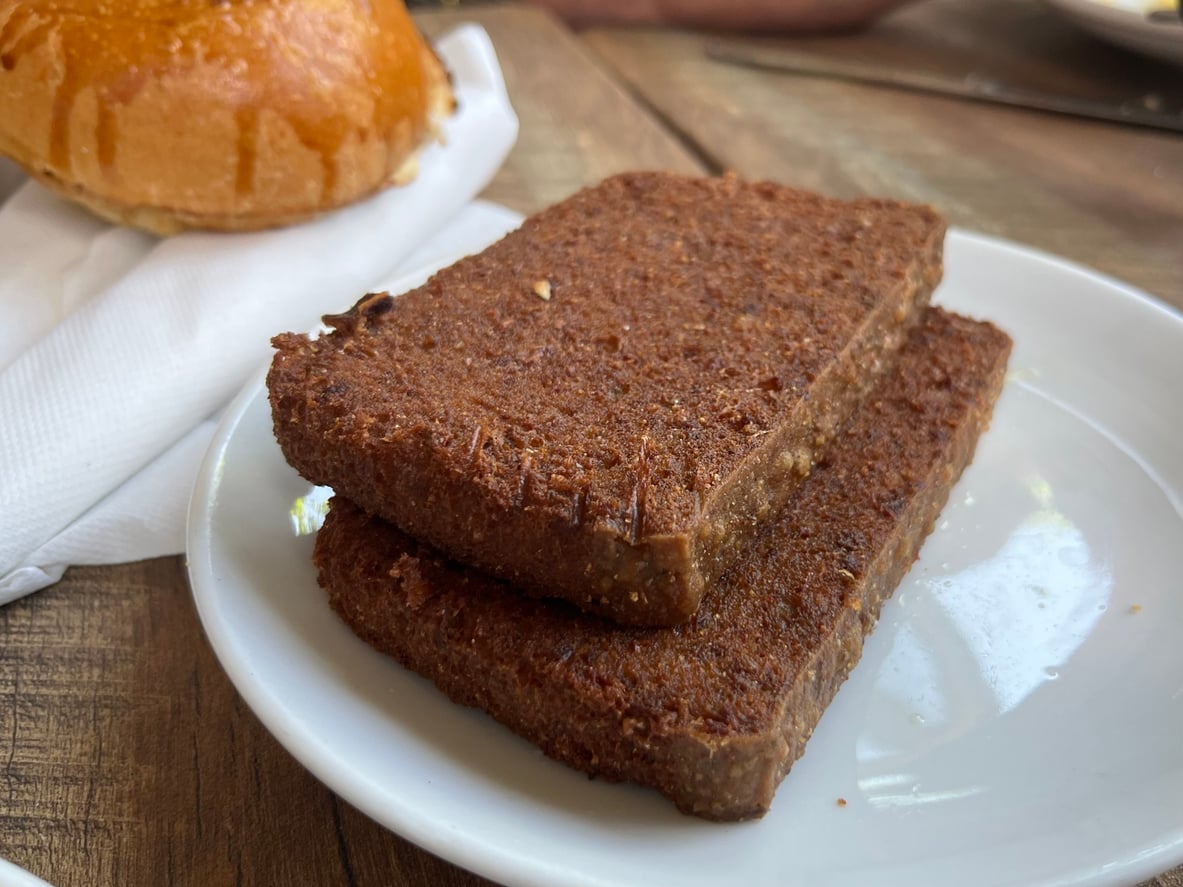 Two slices of dense, dark brown bread are stacked on a white plate next to a napkin and part of a bun on a wooden table.