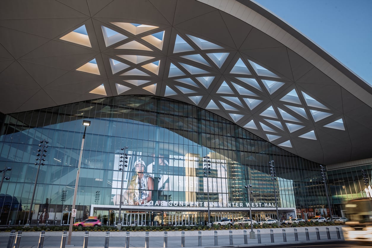 Modern glass facade of the Las Vegas Convention Center West Hall with geometric roof patterns, large windows reflecting the city, and an entrance with signage, captured in daylight.