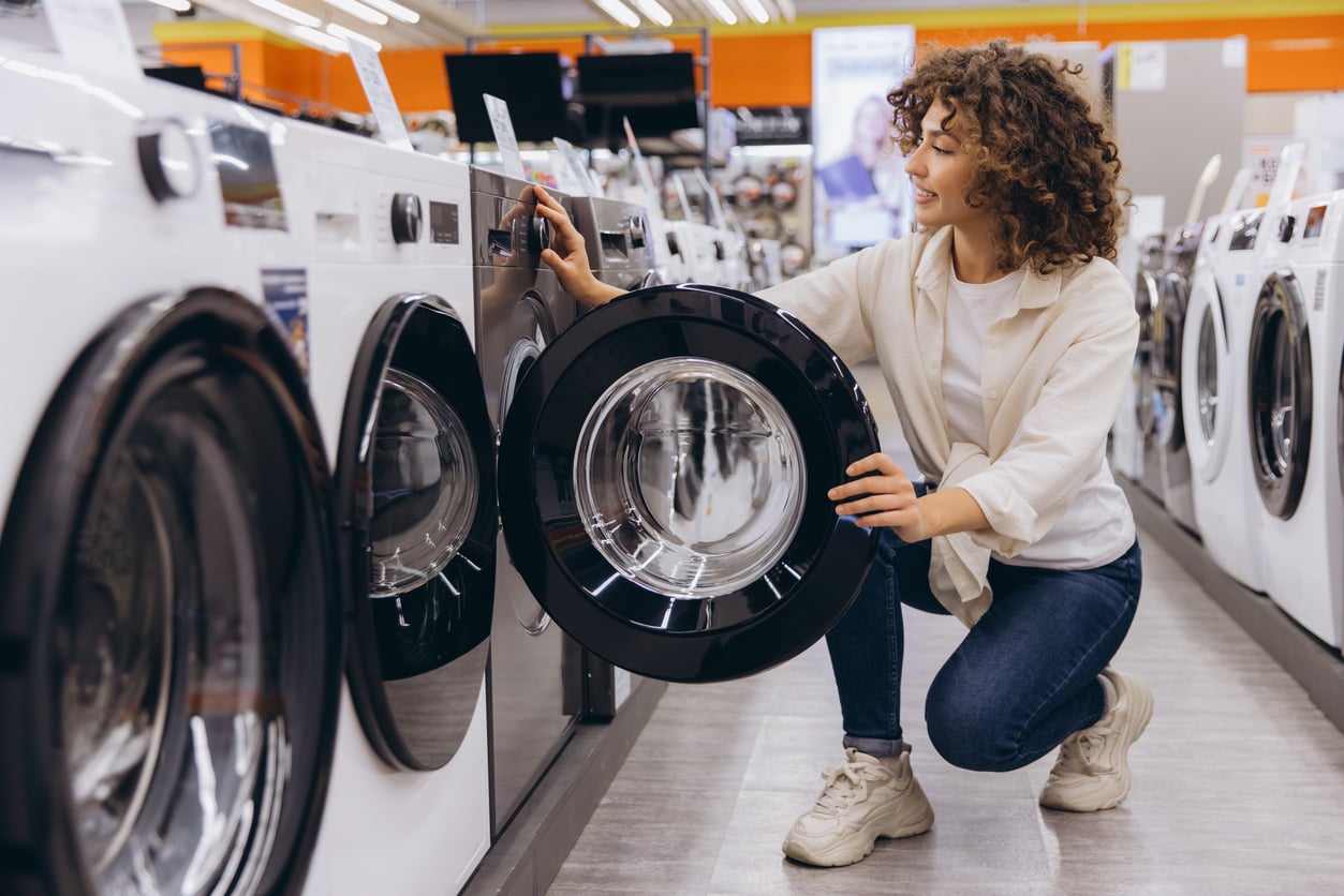 A woman with curly hair kneels in an appliance store, examining the door of a front-loading washing machine among a row of similar machines.