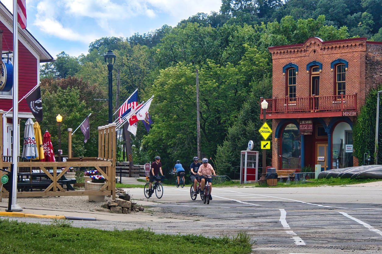 Four people ride bicycles on a small-town street lined with flags, trees, and historic brick buildings, including a museum, on a sunny day.
