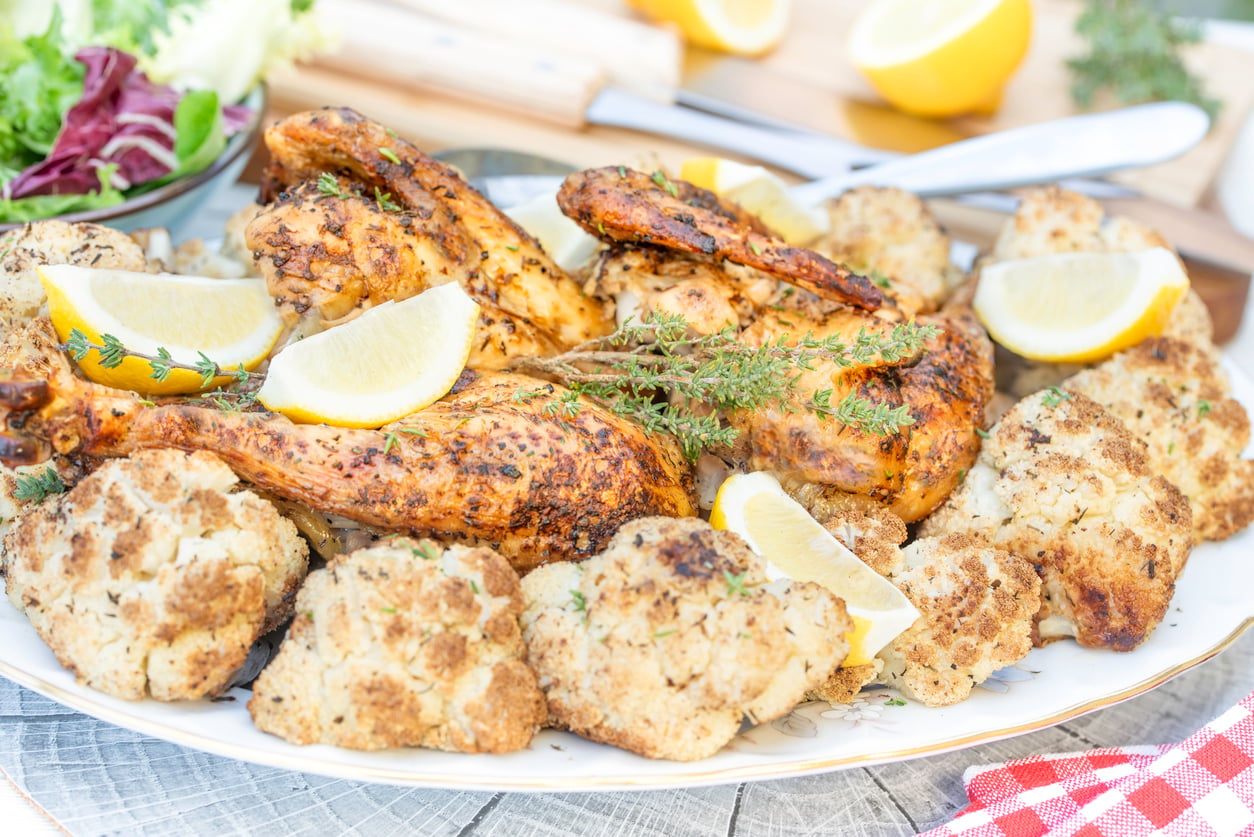 Roasted chicken garnished with lemon wedges and fresh herbs, surrounded by seasoned roasted cauliflower on a white platter, with a salad bowl and lemon halves in the background.