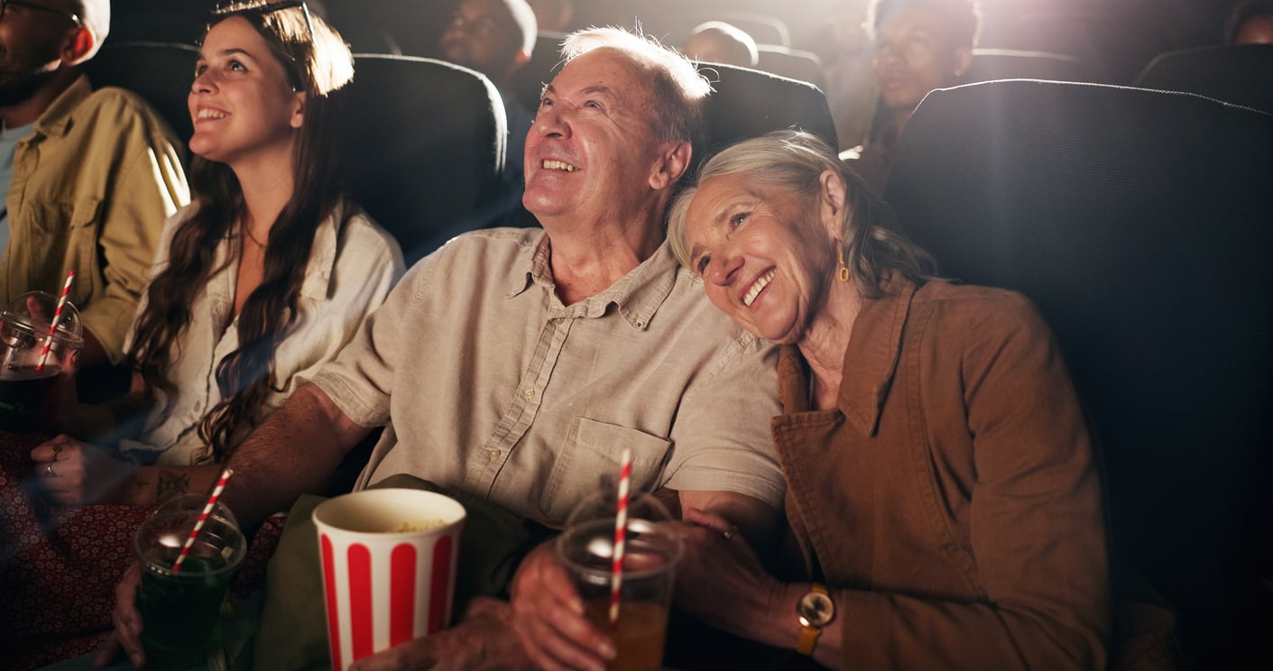 An elderly couple sits closely together, smiling and enjoying drinks and popcorn at a movie theater with other people around them—sharing a simple joy that’s become one of the things families spend less on, yet still brings them closer together.