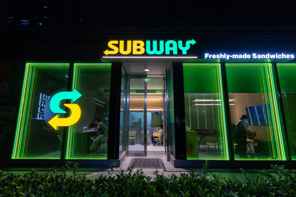 A Subway restaurant entrance at night with bright green and yellow neon lights, displaying the Subway logo and “Freshly-made Sandwiches” sign above the door.