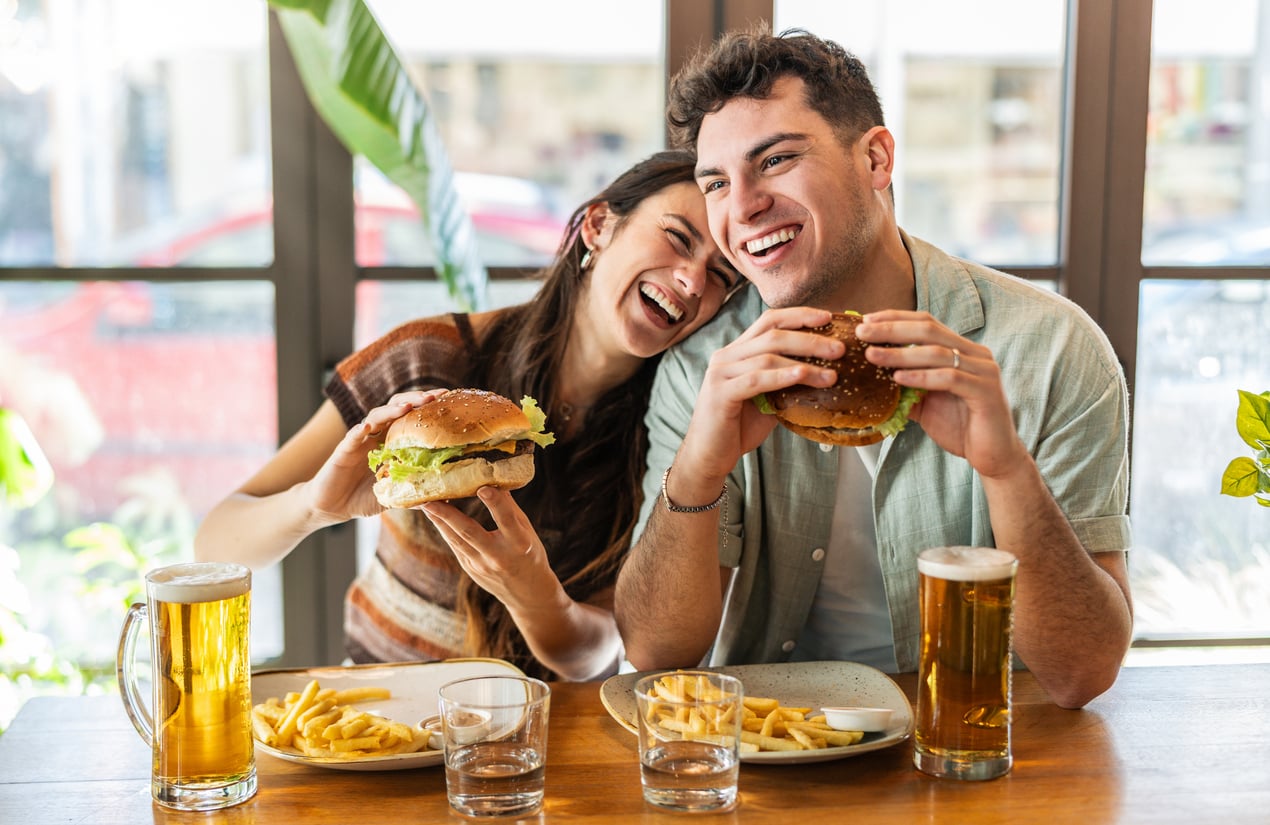 A smiling couple sits at a table, laughing while holding large burgers. Plates of fries, two glasses of beer, and water glasses are on the table in a bright, casual restaurant setting.