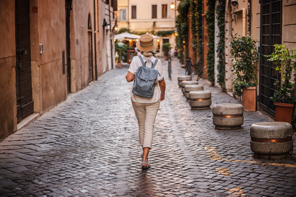 A person wearing a hat and backpack walks alone down a cobblestone street lined with plants and buildings in a European city, perhaps reflecting on vacation mistakes while exploring new sights.