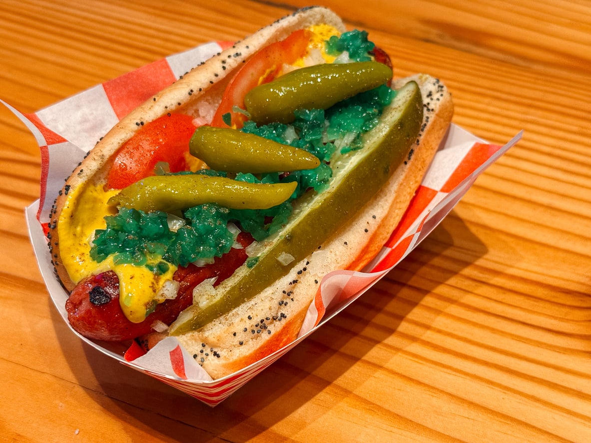 A poppy seed hot dog bun with a sausage, mustard, tomato slice, pickle spear, sport peppers, onions, and green relish on a wooden table in a paper tray.
