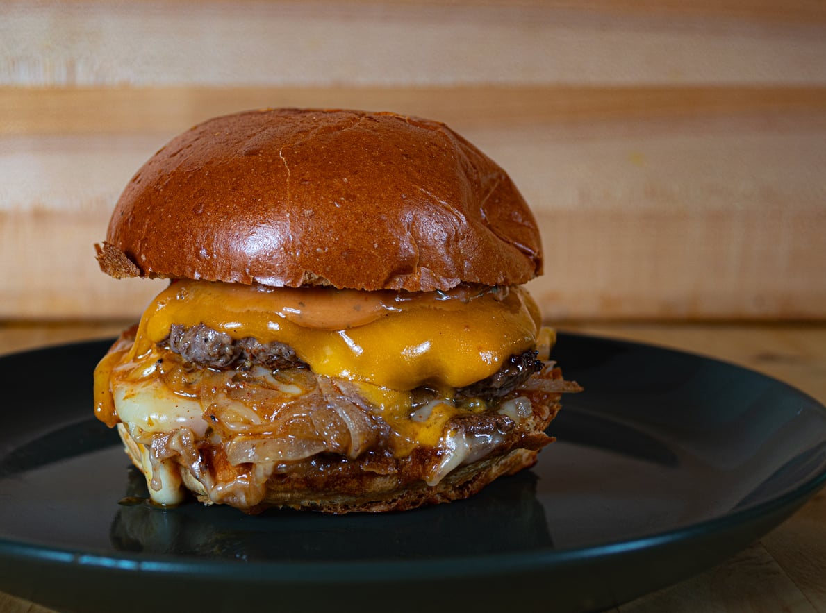 A close-up of a cheeseburger with melted cheese, caramelized onions, and a shiny brioche bun—one of the restaurant specialties—sits on a dark plate against a wooden background.