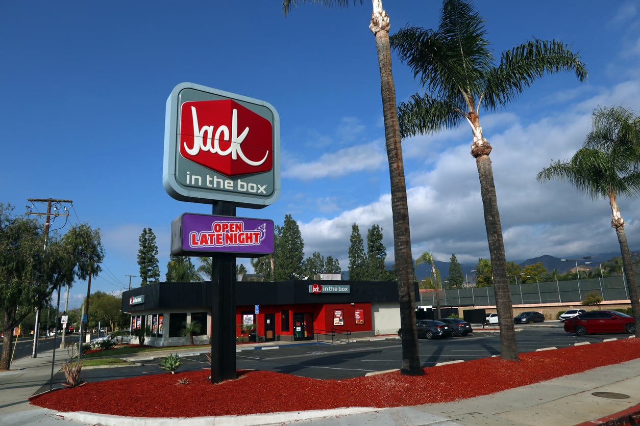 A Jack in the Box fast food restaurant with a large sign displaying the logo and “Open Late Night” under palm trees, with a clear sky and mountain backdrop.