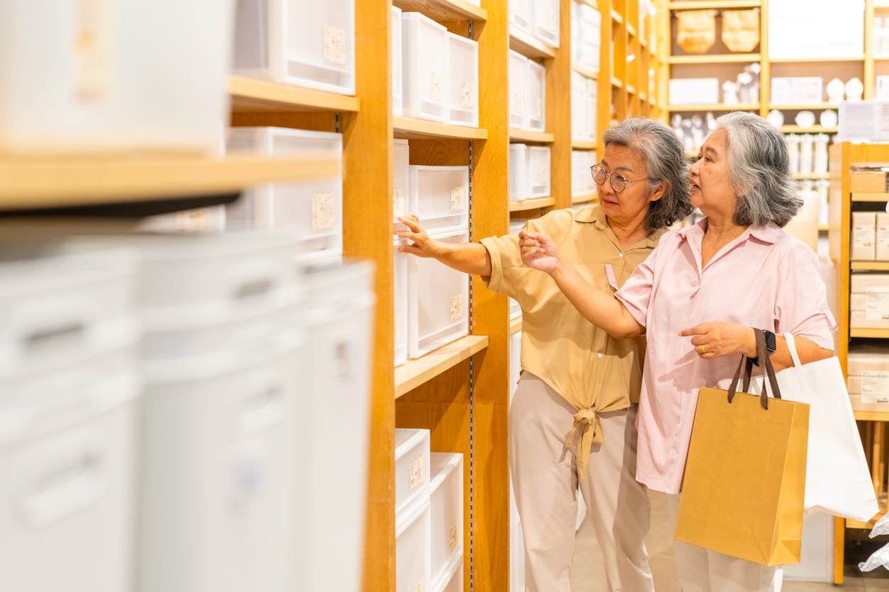 Two older women shop together, looking at storage bins on a store shelf—perhaps considering practical ways to organize things families spend less on. One holds a shopping bag while the other reaches toward the shelves, both appearing engaged and focused. Bright lighting and wooden shelves fill the background.