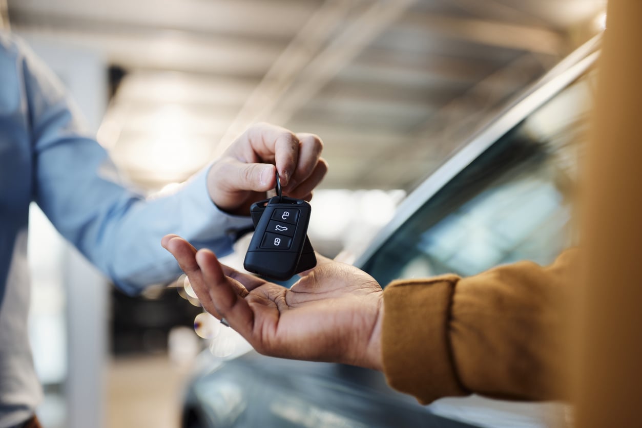A person hands over a car key fob to another person, with a car in the background, suggesting a car sale, rental, or handover—possibly taking advantage of free Costco membership perks related to auto services.
