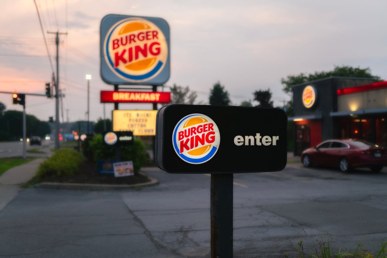 A Burger King “enter” sign is in focus in the drive-thru lane, with the restaurant and a large Burger King sign in the background at dusk. Cars and streetlights are visible.
