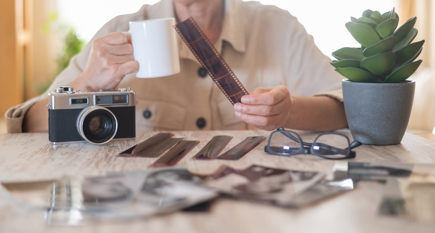 A person holds a mug and a strip of photo negatives at a table with a vintage camera, black glasses, a potted plant, and scattered photographs and negatives.