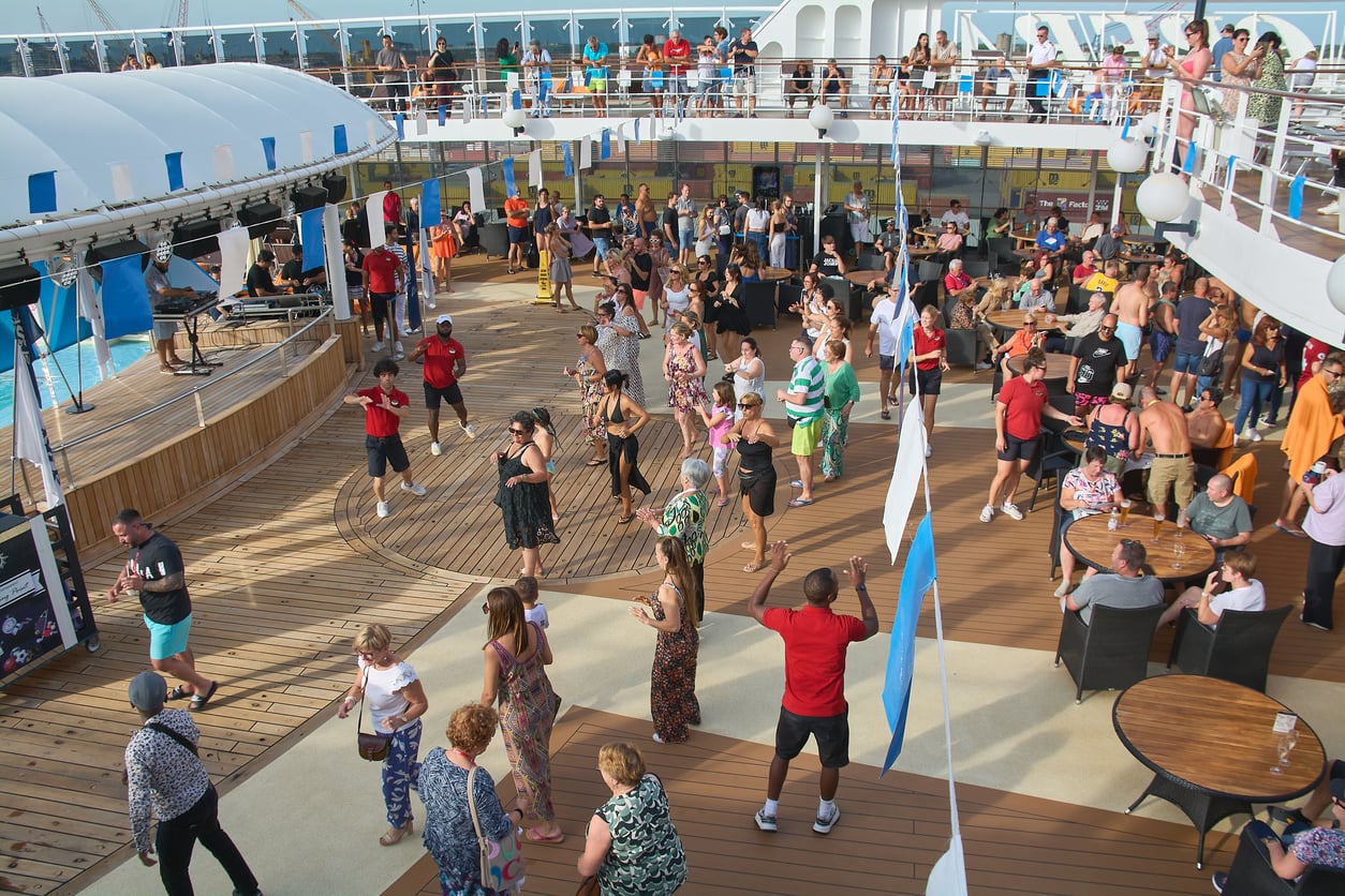 People participate in a group dance activity on a cruise ship deck near a pool, with onlookers and staff nearby under sunny weather.