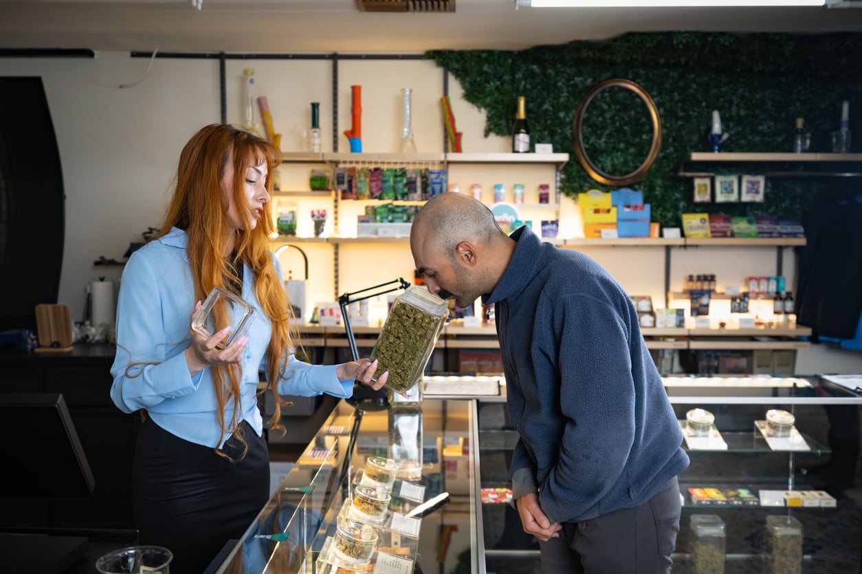 A woman shows a jar of cannabis to a man who is smelling it inside a modern dispensary, with shelves of products visible in the background.