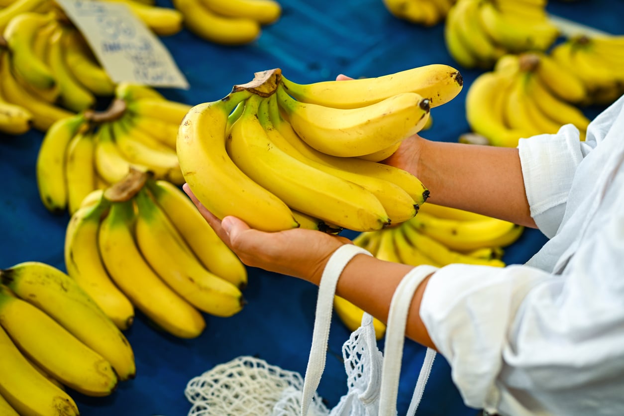 A person holds a bunch of ripe yellow bananas at a market—one of those everyday foods that used to be considered fancy—with more bananas displayed on a blue table in the background.
