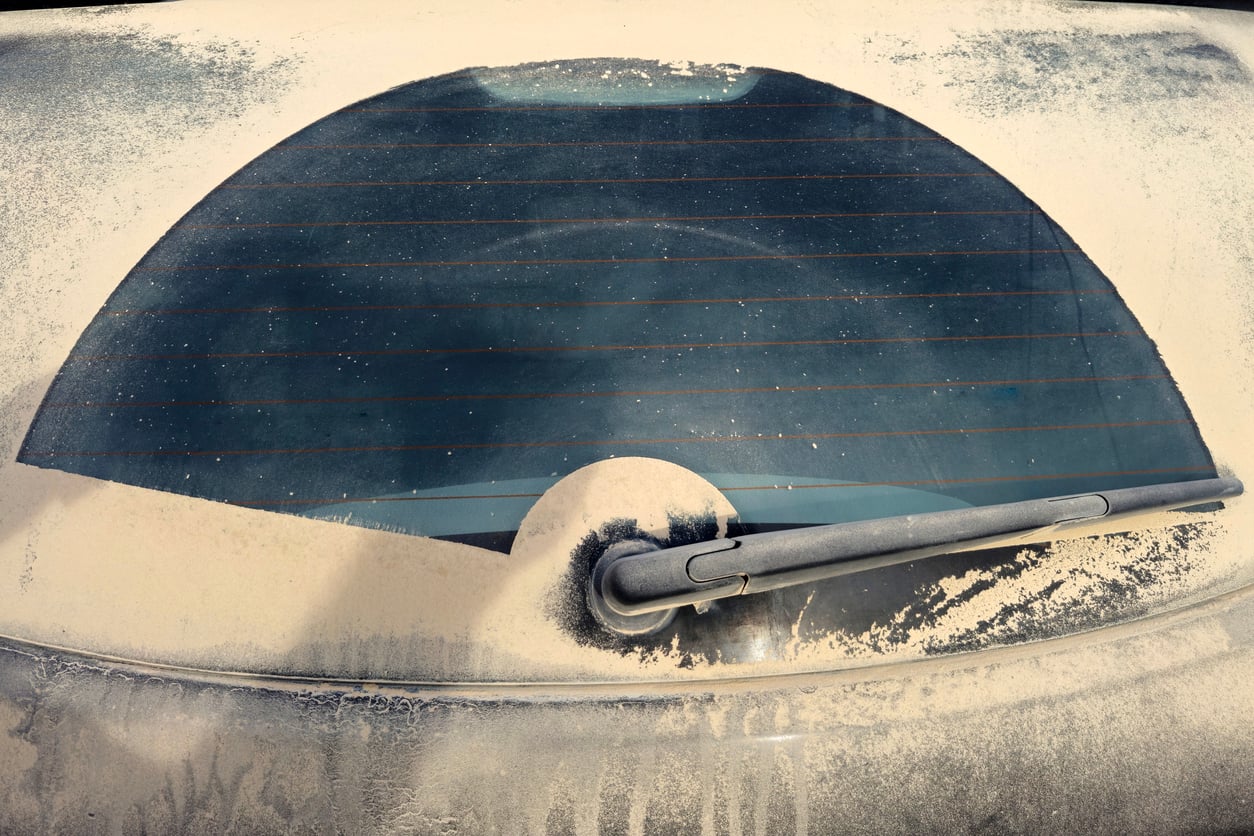 A dusty car rear window with a windshield wiper has cleared a semicircle, leaving a clean area among the dirt. Horizontal defrost lines are visible across the glass.