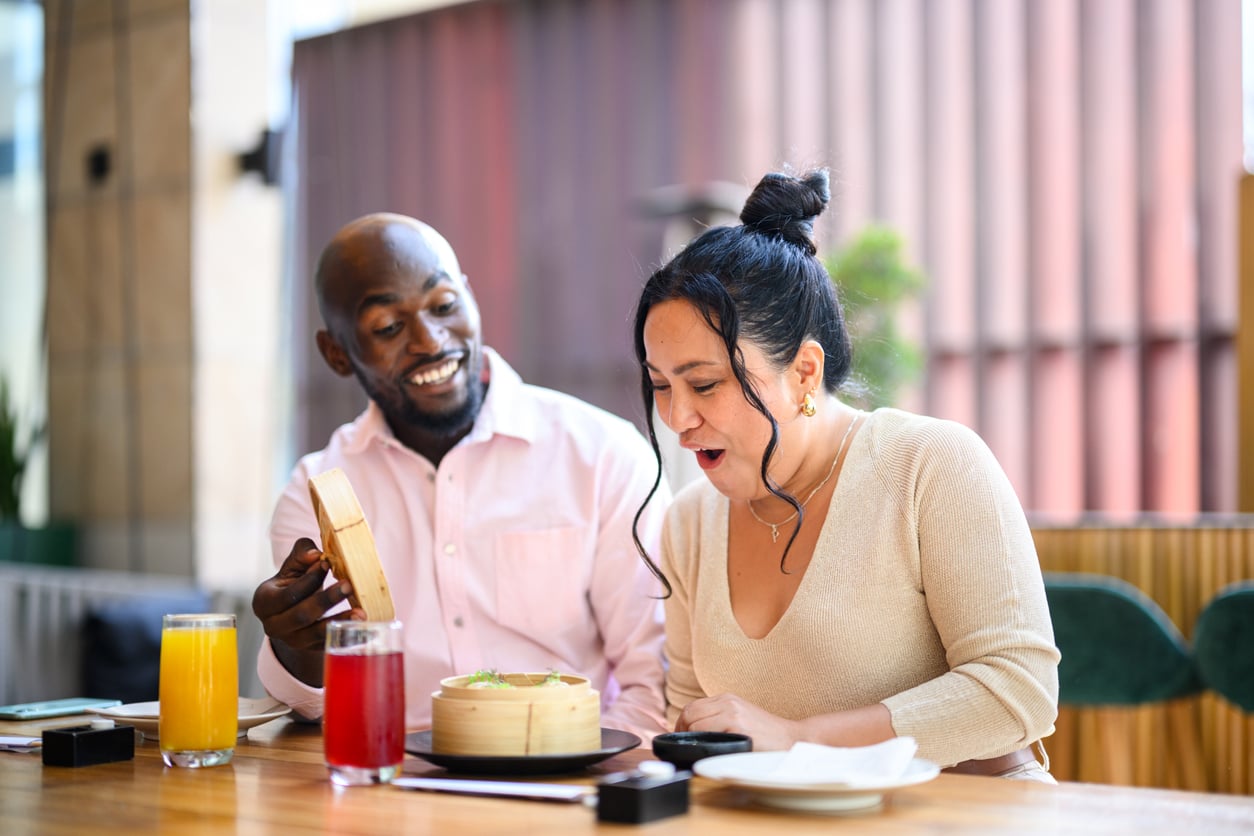 A man and woman sit at an outdoor table, smiling and surprised as they open a bamboo steamer with food inside. There are orange and red drinks in front of them.