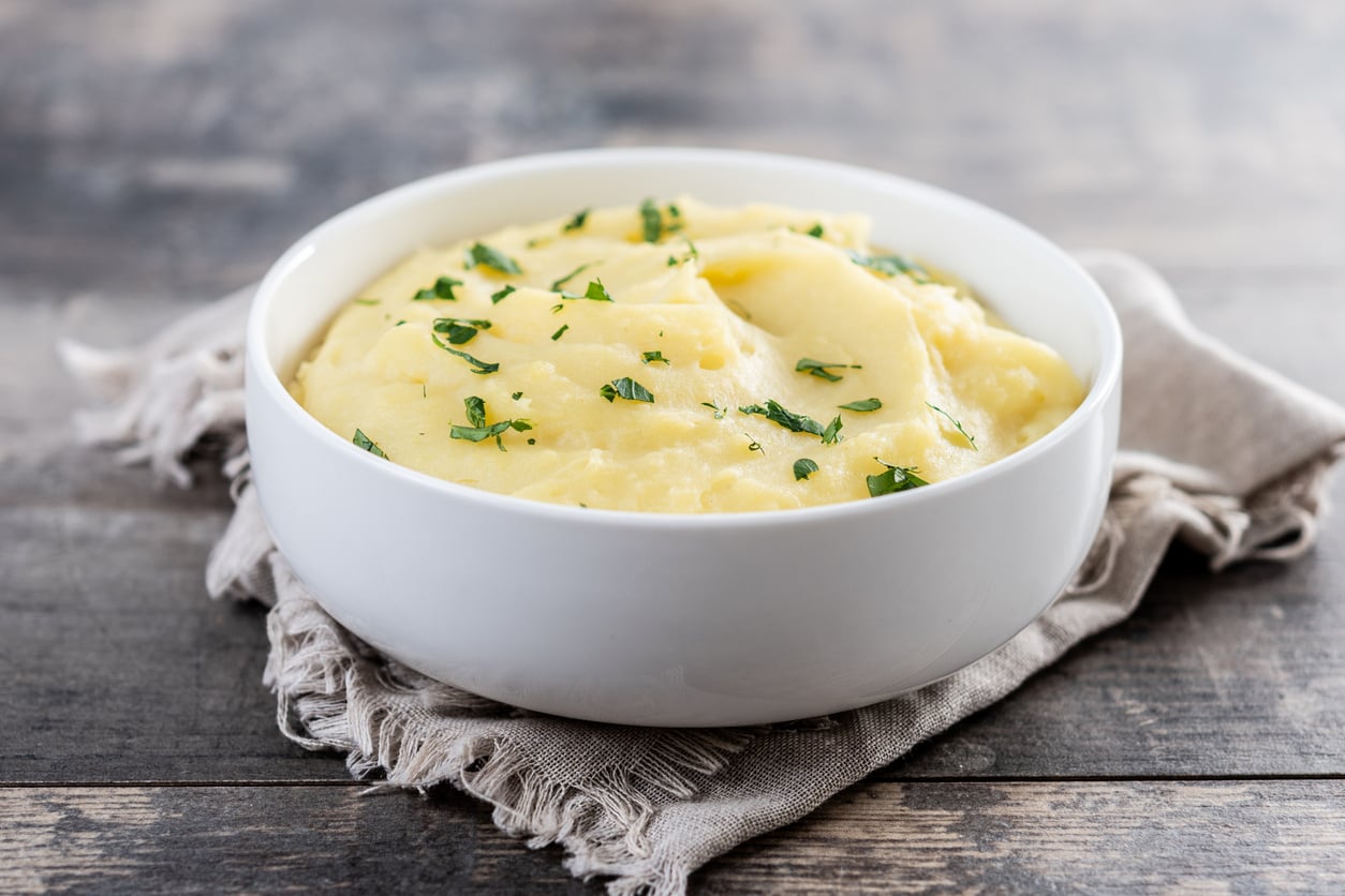 A white bowl filled with creamy mashed potatoes garnished with chopped herbs sits on a cloth napkin atop a rustic wooden table.