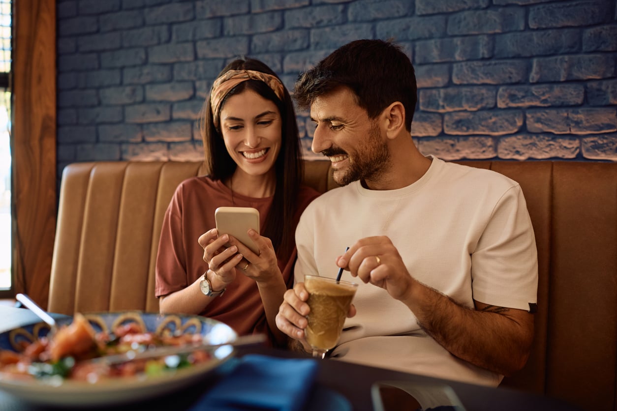 A smiling couple sits in a booth at a restaurant. The woman shows something on her phone to the man, who holds an iced coffee. A plate of food is on the table in the foreground.