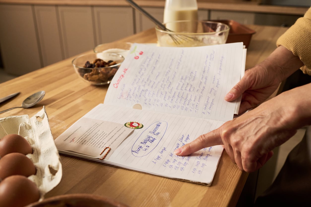 A person points at a handwritten recipe in an open notebook on a kitchen counter, surrounded by eggs, a glass bowl, a spoon, and a carton of milk.