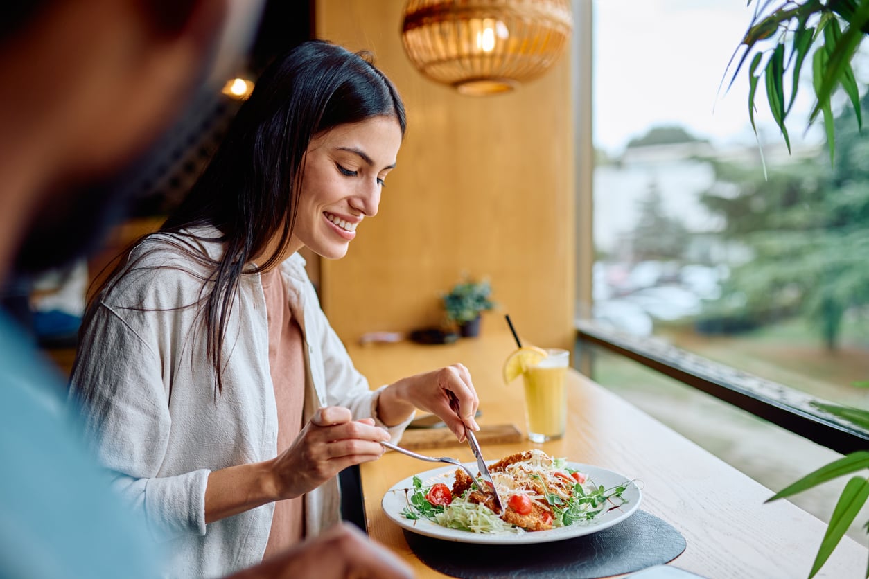 A woman smiles while eating a meal at a restaurant table by a window. She holds a fork and knife over a plate of salad; a drink with a lemon slice sits nearby. Warm light fills the cozy indoor setting.
