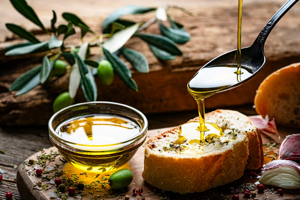 A spoon pours olive oil onto a slice of bread topped with herbs. Nearby are a bowl of olive oil, olives, garlic cloves, and green leaves on a rustic wooden surface—an inviting display of everyday foods that used to be considered fancy.