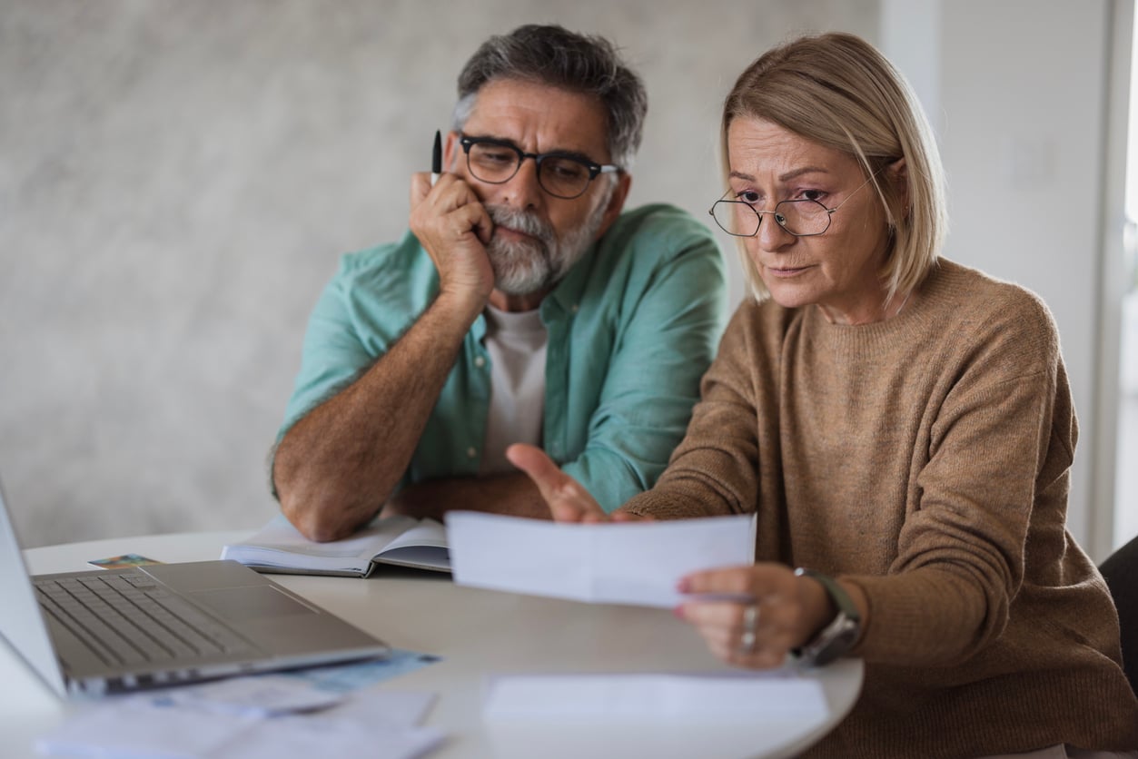 An older couple sits at a table looking concerned while reviewing paperwork in front of a laptop, suggesting they are dealing with finances or bills.
