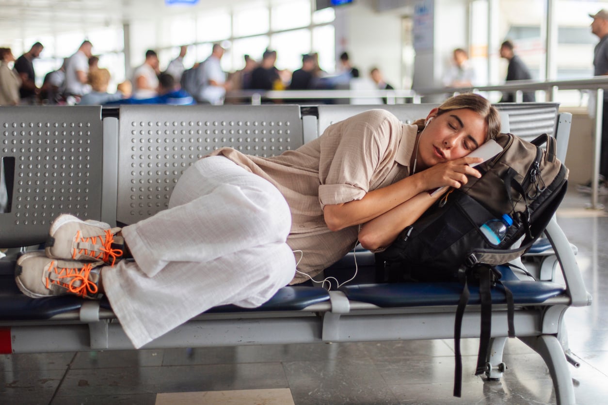 A woman in light clothing sleeps on airport seats, using her backpack as a pillow. Other travelers are in the background.