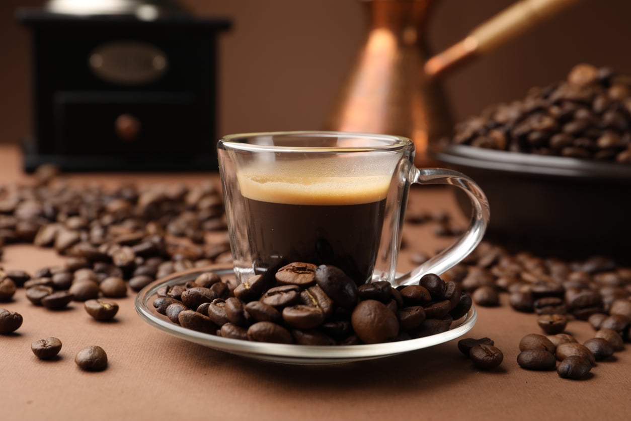 A clear glass cup of espresso sits on a saucer surrounded by coffee beans, with more beans scattered around and coffee-making equipment blurred in the background—a simple yet elegant reminder of how espresso once joined the ranks of everyday foods that used to be considered fancy.