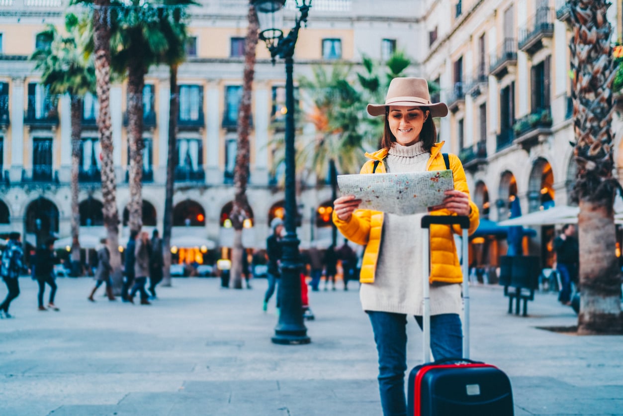 A woman in a yellow jacket and hat stands in a plaza, holding a map and a suitcase—perhaps reflecting on common vacation mistakes. People and palm trees are in the background near tall buildings with balconies.