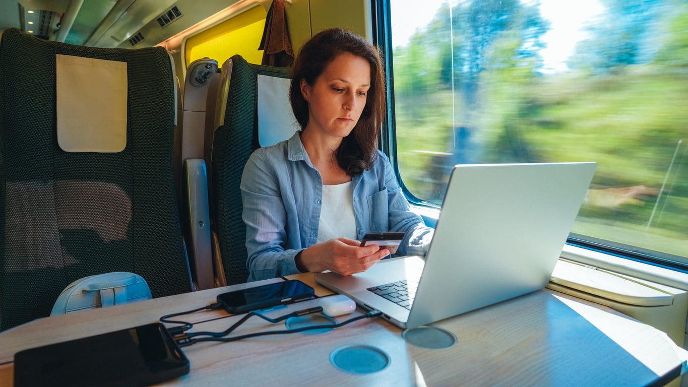 Woman working on a laptop and using her phone at a table on a train, with various electronic devices on the table and blurred green scenery visible outside the train window.