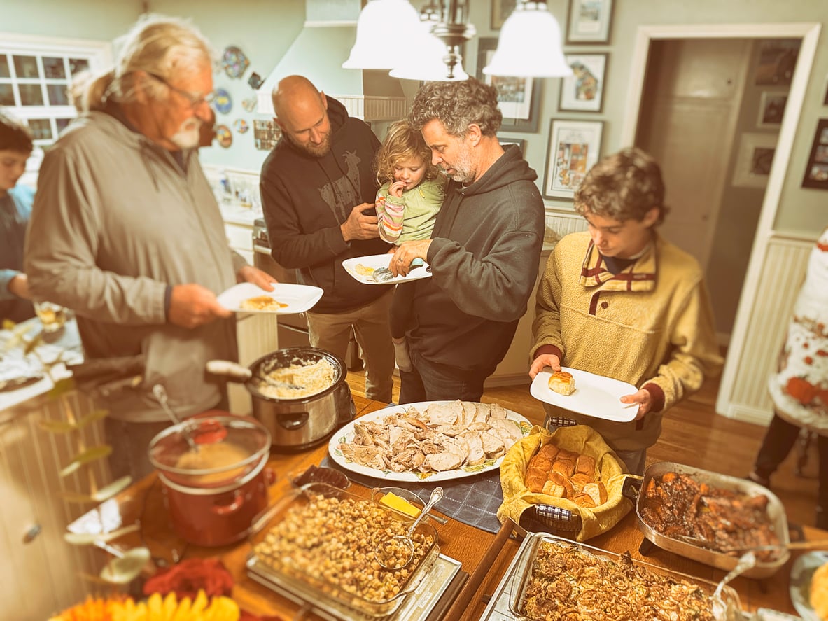 People stand around a kitchen island filled with various dishes, serving themselves food. A man holds a young child while others fill their plates, suggesting a family gathering or holiday meal. The atmosphere appears warm and lively.