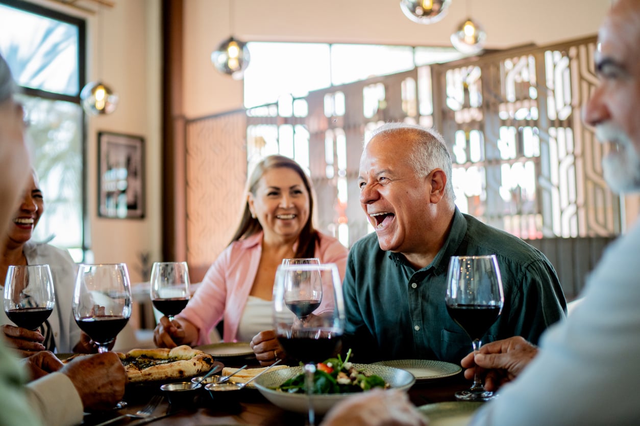 A group of older adults sit at a restaurant table, laughing and enjoying glasses of red wine and plates of food in a bright, modern setting.