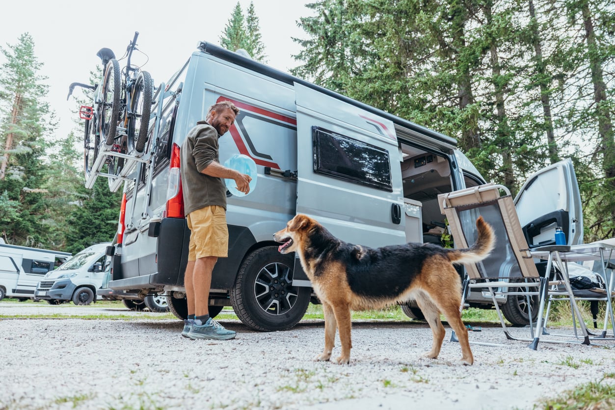 A man stands by a camper van in a forested campsite, holding a water bottle. A black and brown dog stands near him. Two bicycles are mounted on the van. Outdoor chairs and a table are set up beside the van.