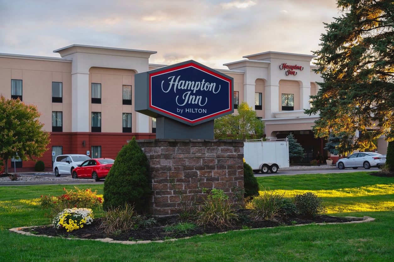 A Hampton Inn by Hilton hotel, part of renowned hotel chains, with a large sign in front, is surrounded by grass, flowers, trees, a red car, and a white trailer in the parking lot. The building is beige with multiple windows.